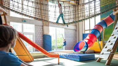 In einer Halle stehen Rutschbahnen, eine Hängebrücke und dicke Matten Spieltürme aus Holz. Am Boden liegen Matten und die Türme werden von einer Hängebrücke verbunden.