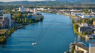 Luftaufnahme des Rheins mit Booten, umgeben von Stadtgebäuden und Bergen im Hintergrund.