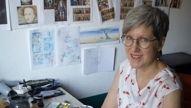 Person mit Brille sitzt an einem Schreibtisch, umgeben von Zeichnungen an der Wand.