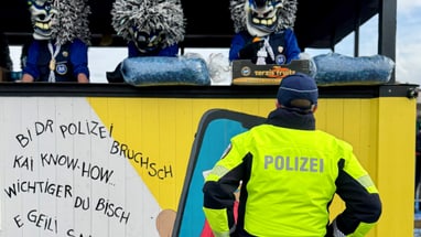 Polizist betrachtet verkleidete Personen auf einem Balkon mit Graffiti-Wand darunter.