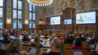 Menschen in einem Saal mit Holzwänden, zuhören einem Vortrag.