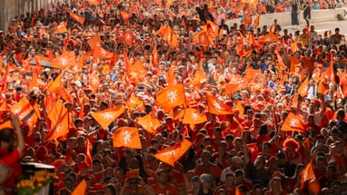Menschenmenge mit orangefarbenen Fahnen bei einer Veranstaltung im Freien.
