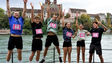 Sechs junge Erwachsene, die am Quer durch Basel teilnehmen, springen in die Luft, um für das Foto zu posen. Alle halten eine Stafette in der Hand. Im Hintergrund sieht man den Rhein und hintendran das Münster. 