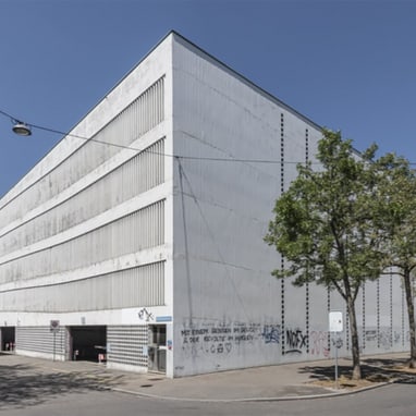 Weisses Parkhaus mit Graffiti an der Strassenecke, umgeben von Bäumen.