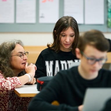 Michèle Minelli spricht mit Schüler am Tisch, weiterer Schüler im Vordergrund mit Laptop.