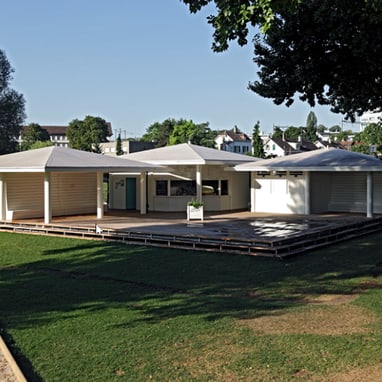 Moderner Pavillon im Grünen mit Bäumen im Hintergrund.",