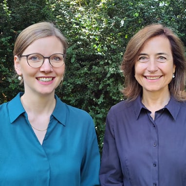 Zwei lächelnde Frauen vor Büschen im Freien.