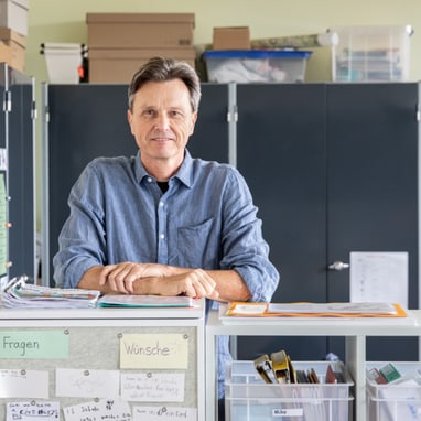 Person in blauem Hemd steht vor einem Schreibtisch mit Regalen und Büromaterial.