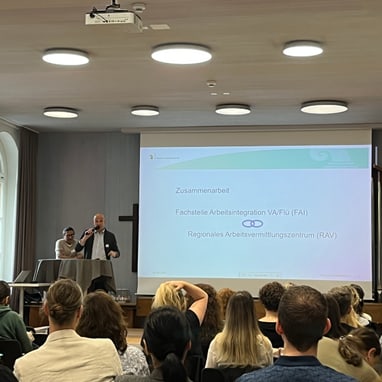 Mann hält Präsentation vor Publikum in einem Raum mit Projektor.