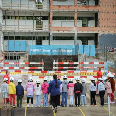 Kinder mit Helmen bei einer Baustellenbesichtigung vor einem Gebäude im Bau.