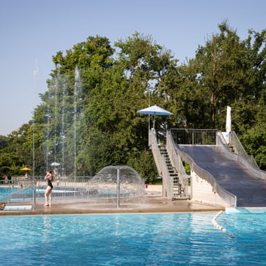 Öffentliches Freibad mit Wasserrutsche und Fontänen.