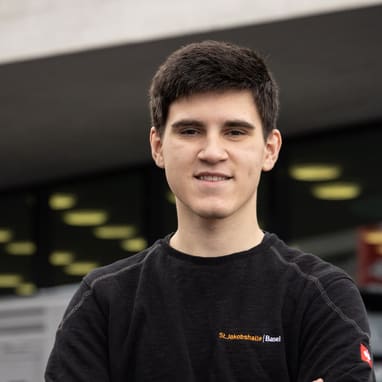 Mann vor Gebäude mit grossen Fenstern, trägt schwarzen Pullover.