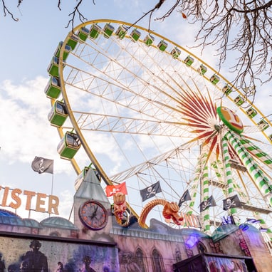 Riesenrad und Geisterbahn auf Jahrmarkt mit Baumzweigen im Vordergrund.