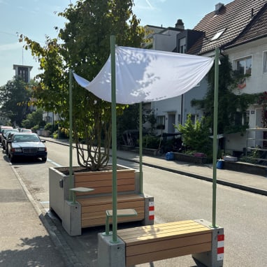 Holzbänke mit Sonnensegel neben Baum auf Stadtstrasse.