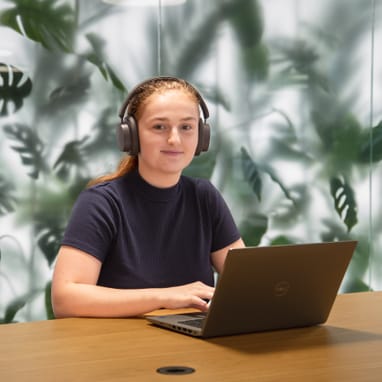 Frau mit Kopfhörern arbeitet am Laptop, Pflanzenhintergrund.",