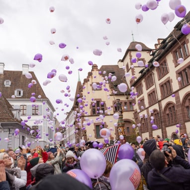 Menschenmenge mit lila Luftballons in historischer Stadtumgebung.