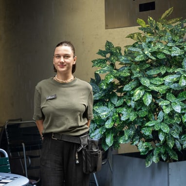 Die Restaurationsfachfrau Manon Furler lächelt im Freien neben Pflanze vor Nomad-Schild.