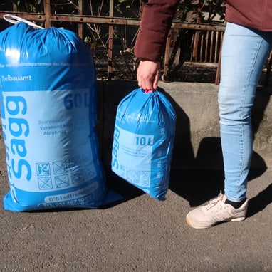 Person hält blauen Abfallsack neben grösserem Sack auf Gehweg.