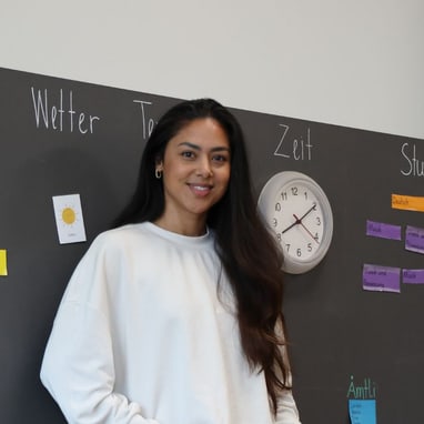 Frau vor Tafel mit Uhr und Beschriftungen in Klassenzimmer.