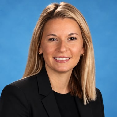 Person mit blonden Haaren in schwarzem Blazer vor blauem Hintergrund.