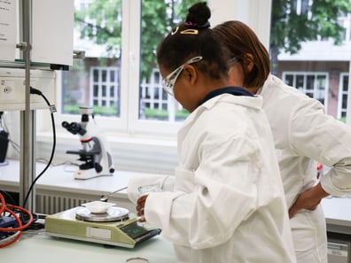 Zwei Personen im Labor mit Waage und Mikroskop im Hintergrund.