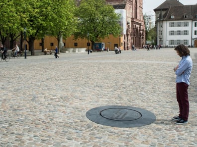 Person steht auf dem Platz vor dem Basler Münster und betrachtet die kreisrunde Gusseisenplatte im Plastersteinboden, welche den römischen Sodbrunnen markiert