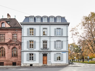 Drei-stöckiges hellgraues Gebäude an einer Strasse, Herbstbäume im Hintergrund.