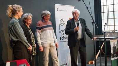 Gruppe von Menschen auf einer Bühne mit Mikrofon und Banner im Hintergrund.
