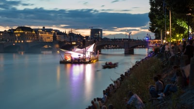 Menschen am Flussufer mit beleuchtetem Floss bei Nacht.