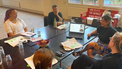 Personen in Besprechungssaal mit Laptop auf dem Tisch, Fenster im Hintergrund.