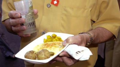 Person in Hemd hält in einer Hand einen Teller mit Raclette und in der anderen einen Mehrwegbecher.