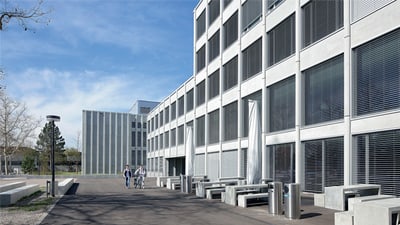 Fassade des Schulhauses, davor stehen einige Tische und Bänke aus Beton.