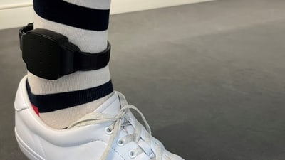 Weisse Turnschuhe und elektronische Fussfessel am Bein mit gestreiften Socken.