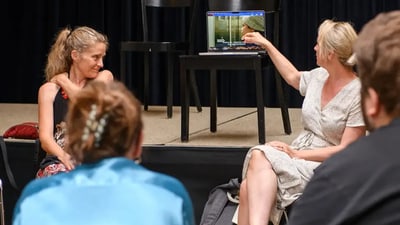 Frauen bei einer Präsentation, eine zeigt auf den Laptopbildschirm, sitzend vor einer Bühne 