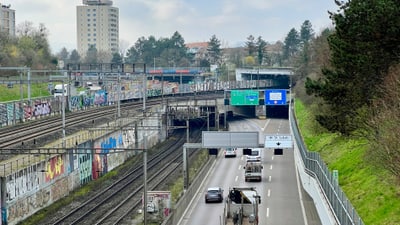 Blick auf Autobahn mit Autos neben Bahngleisen, Gebäude im Hintergrund.