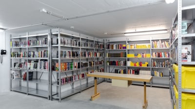 Kellerbibliothek mit Metallregalen, Büchern und Holztisch.