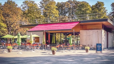 Aussenbereich eines Cafés mit Tischen und Stühlen, umgeben von Bäumen.