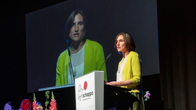 Frau im gelben Cardigan hält Vortrag auf Bühne mit Blumenarrangement.