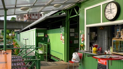 Kiosk-Theke und Tisch mit Stühlen im Vordergrund, dahinter die Umkleiden. Der Kiosk und die Umkleiden sind grün mit weissen Akzenten. 