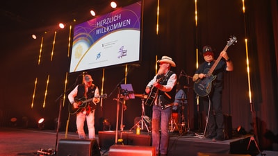 Band spielt auf einer Bühne mit Begrüssungsbildschirm im Hintergrund.
