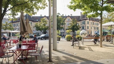 Platz mit Café-Tischen und Bänken, wo Leute sitzen und dahinter Wohnhäuser