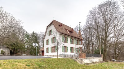 Altes Haus mit Erker. Davor ein Platz mit einem Basketballkorb und dahinter einige Bäume.