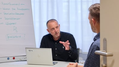 Zwei Männer, die in einem Büro am Tisch sitzen und diskutieren, mit Flipchart daneben.