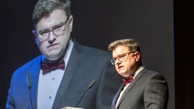 Mann im Anzug hält eine Rede am Podium mit Blumen und Leinwand im Hintergrund.