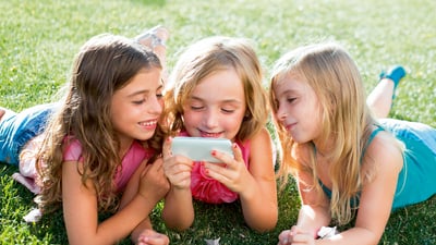 Lächelnde Kinder liegen auf dem Gras und schauen auf ein Smartphone.