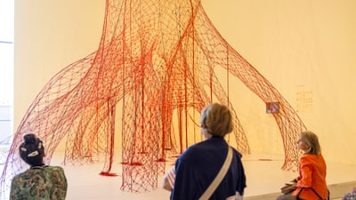 Drei Personen sehen sich eine grosse rote Netzskulptur in einem Raum an.