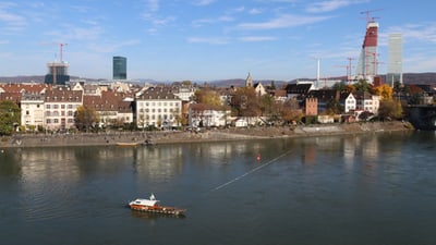 Blick auf Basels Flussufer mit Fähre und Baustellenkräne.