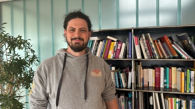 Person in grauem Hoodie vor Bücherregal und Pflanze.