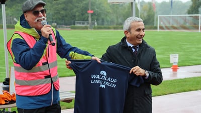 Speaker Markus Harzenmoser und RR Mustafa Atici präsentieren ein Laufshirt auf einem Sportfeld.