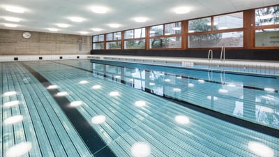 Schwimmbecken auf dessen Wasseroberfläche sich die Deckenbeleuchtung spiegelt. Rechts davon die Fensterfront und dahinter eine Wand mit einer Uhr.